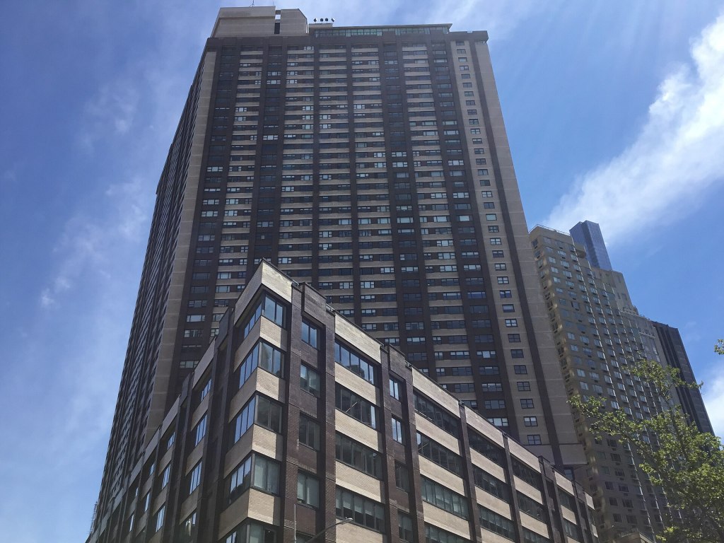 One Lincoln Plaza, the dark brick residential tower at 1900 Broadway across from Lincoln Center, seen from the street against a blue sky